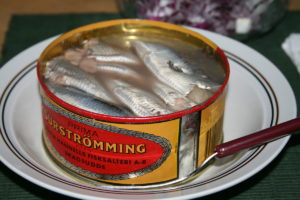 An opened tin of Prima surströmming with fermented herring fillets in brine, on a plate with chopped red onion behind